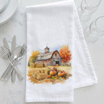 Load image into Gallery viewer, A white kitchen towel with a watercolor autumn barn scene printed on it, styled on a white marble surface with silverware, plates, and wine glasses.
