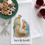 Load image into Gallery viewer, A white kitchen towel with a watercolor illustration of the Holy Family and the text 'Joy to the World' at the bottom, styled on a marble surface with holiday cookies, red mugs, and silverware.
