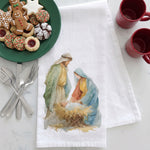 Load image into Gallery viewer, A white kitchen tea towel with a watercolor-style Nativity scene printed on it, laid flat on a light marble surface. A green plate of holiday cookies sits at the top left, silver utensils at the lower left, and two red mugs at the upper right.
