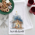 Load image into Gallery viewer, A white kitchen towel with a watercolor-style Nativity scene and the text 'Joy to the World' is laid on a light marble surface. A green plate of cookies, silver forks and spoons, and two red mugs are arranged around it as styled props.
