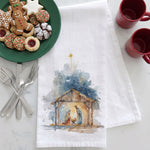 Load image into Gallery viewer, A white kitchen towel with a watercolor nativity scene printed on it, styled on a white marble surface with a green plate of cookies, silverware, red mugs, and the towel.
