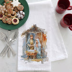 Load image into Gallery viewer, A white kitchen tea towel with a watercolor nativity scene print, styled on a white marble surface with holiday cookies on a green plate, silverware, and red mugs.

