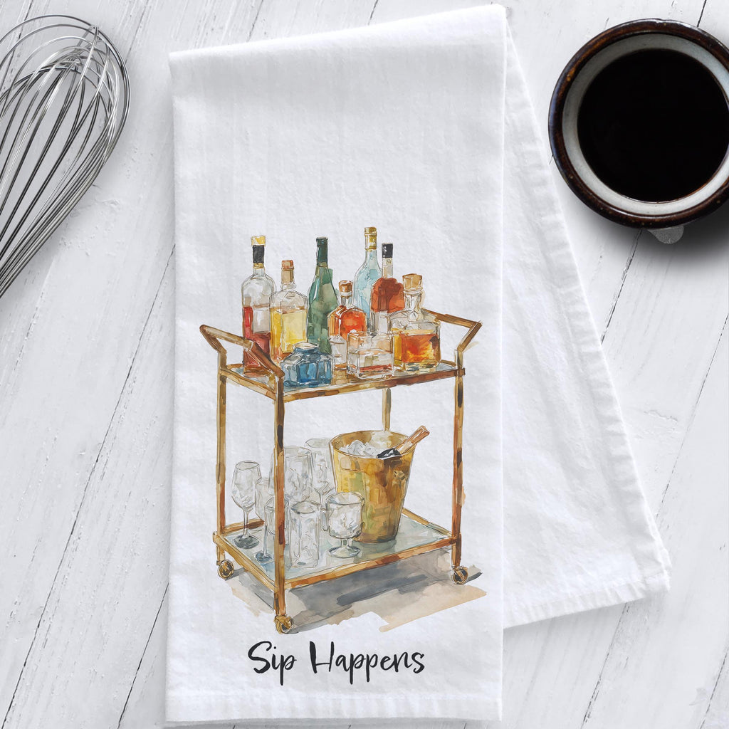 White cotton flour sack tea towel featuring watercolor artwork of a brass bar cart with bottles, glasses, and ice bucket, labeled “Sip Happens.”