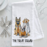 Load image into Gallery viewer, A white kitchen towel with a printed illustration of three dogs dressed in Halloween attire, with the text 'THE TREAT SQUAD' at the bottom. The towel is styled in a flat-lay on a white marble surface with silver cutlery, glassware, and white plates.

