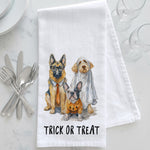 Load image into Gallery viewer, A white kitchen towel with a watercolor illustration of three dogs dressed in Halloween costumes, with the text 'TRICK OR TREAT' printed below. The towel is styled in a flat-lay on a white marble surface with silver cutlery, glassware, and white plates.
