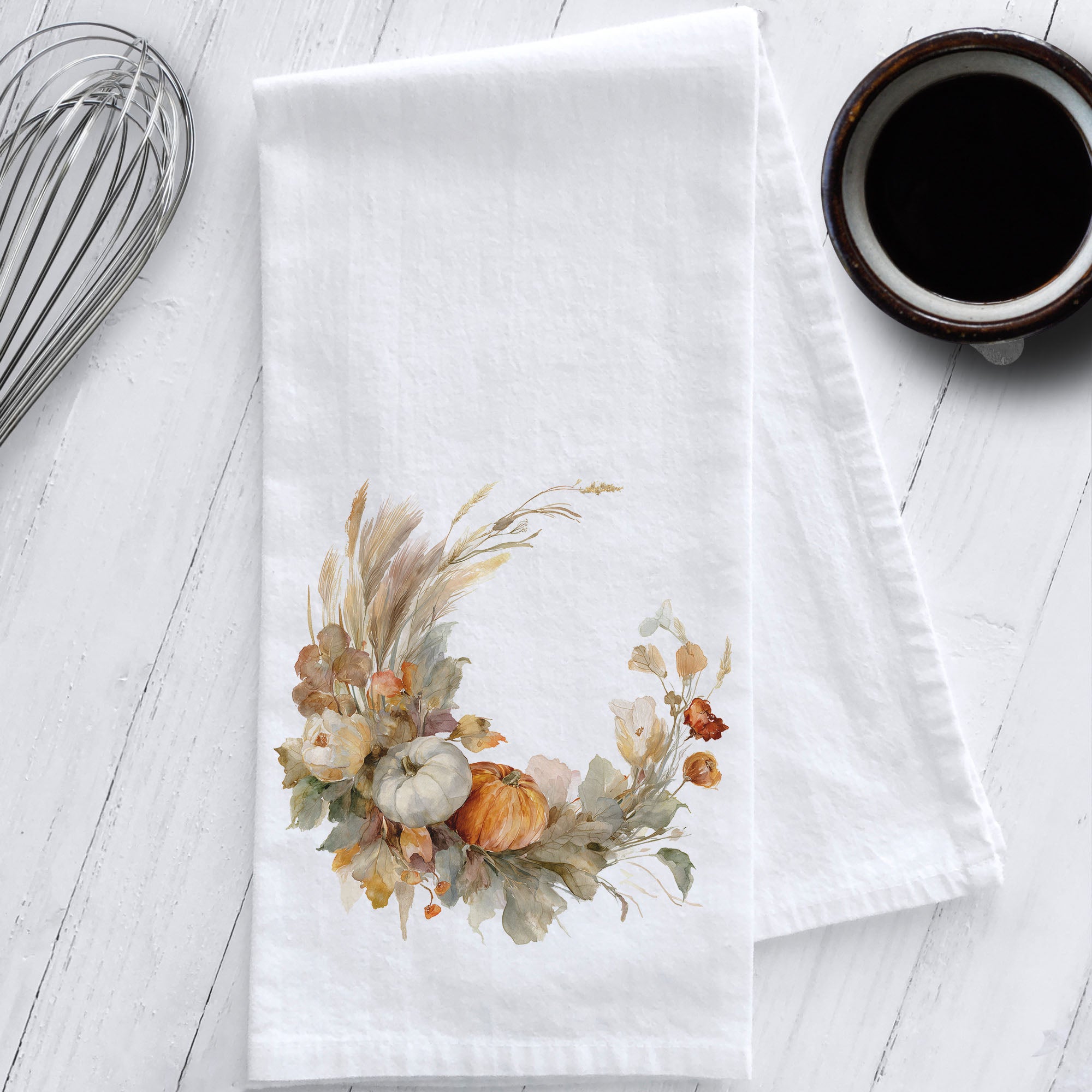 White cotton flour sack tea towel featuring a watercolor boho-style pumpkin wreath with pampas grass and fall florals, styled flat on a white wooden surface.